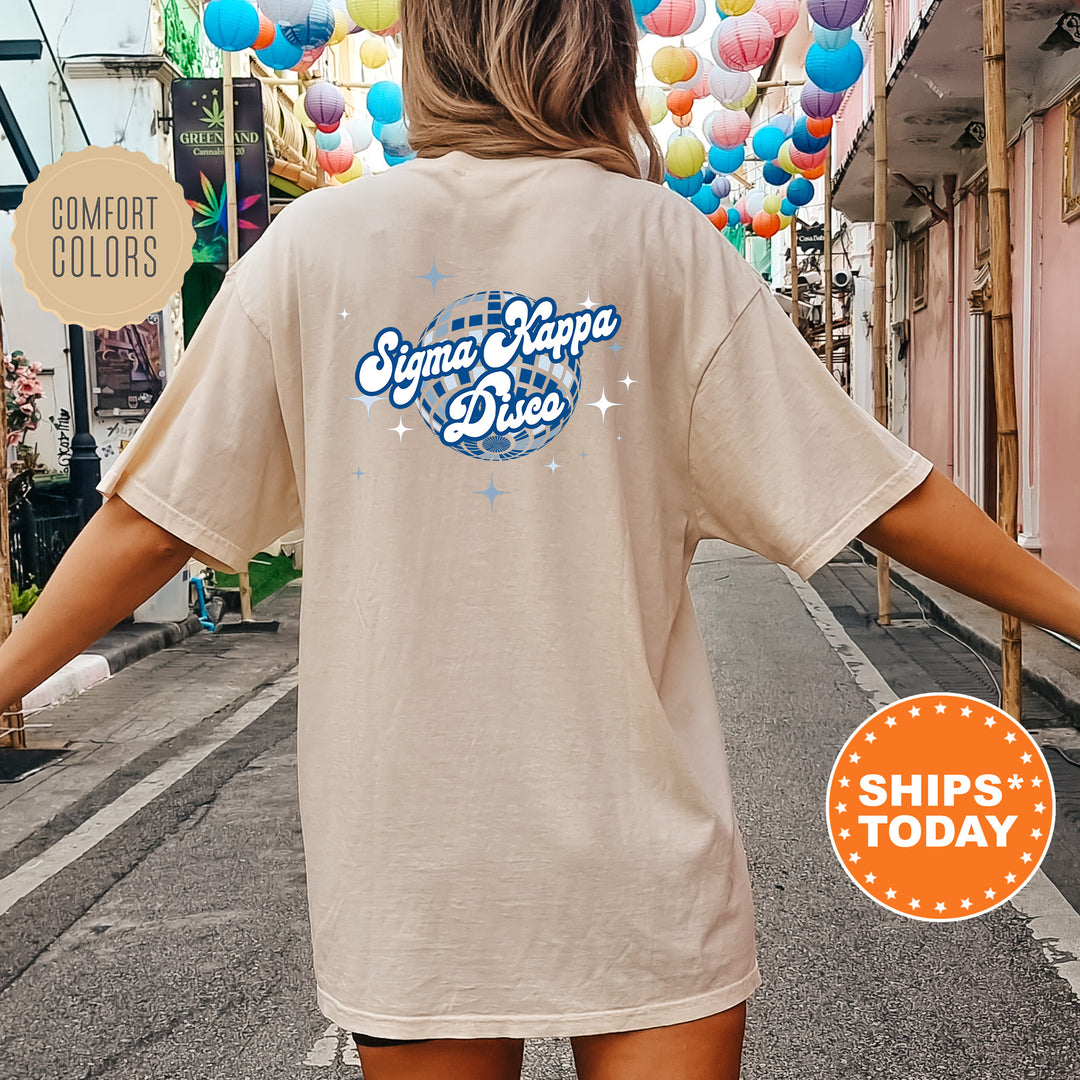 A person wearing a beige t-shirt with a graphic design and text printed on the back, standing on a street with colorful lanterns hanging overhead.