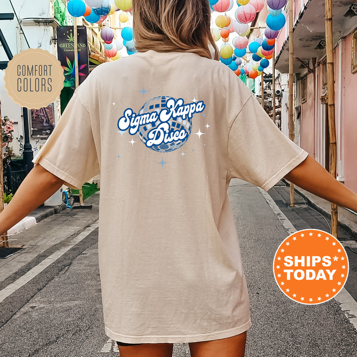 A person wearing a beige t-shirt with a graphic design and text printed on the back, standing on a street with colorful lanterns hanging overhead.