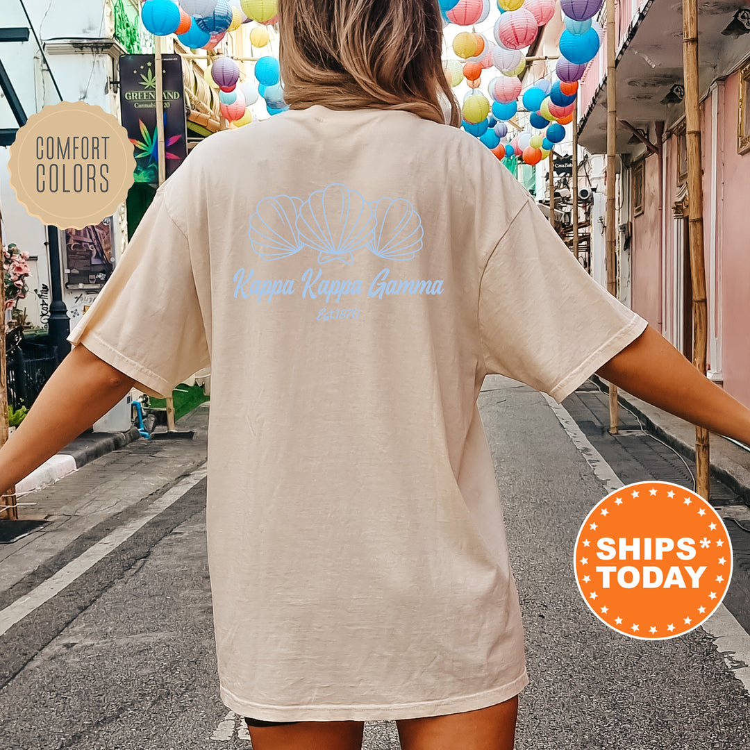 A person wearing a beige t-shirt with a graphic design of seashells and the text "Kappa Kappa Gunna" printed on the back, standing on a street with colorful lanterns hanging overhead.