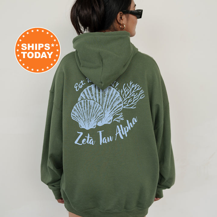 A person wearing a green hoodie with a graphic design of seashells and the text "Zeta Taw Alpha" printed on it.