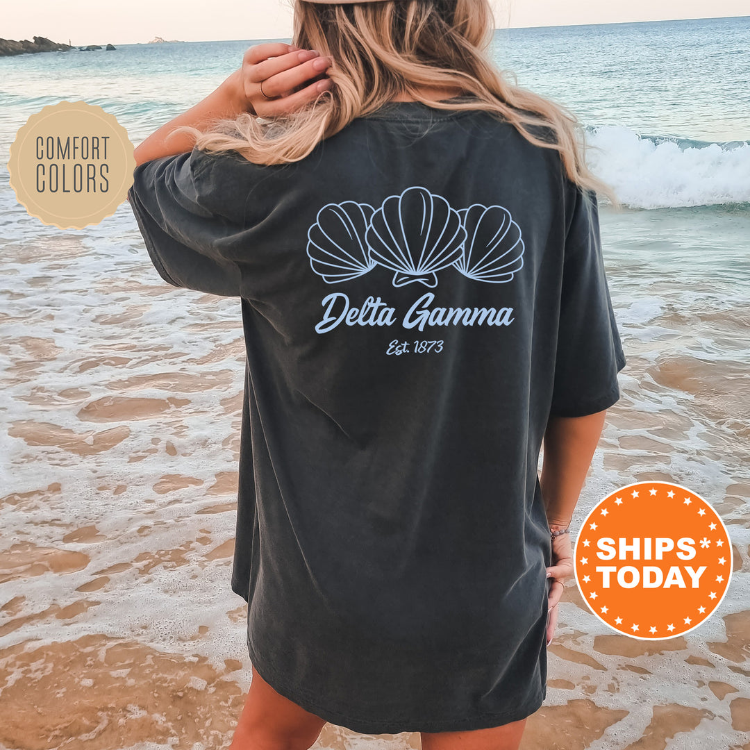 A woman wearing a black t-shirt with a graphic design of three shells on the back, standing on a beach with the ocean in the background.