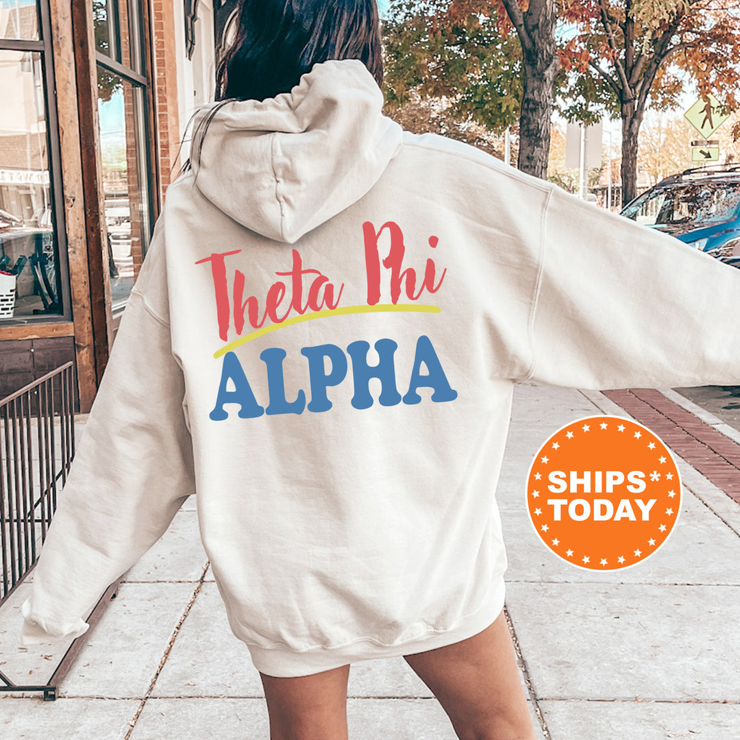 A person wearing a white hoodie with the text "Theta Phi Alpha" printed on it, standing on a sidewalk with trees and a building in the background.