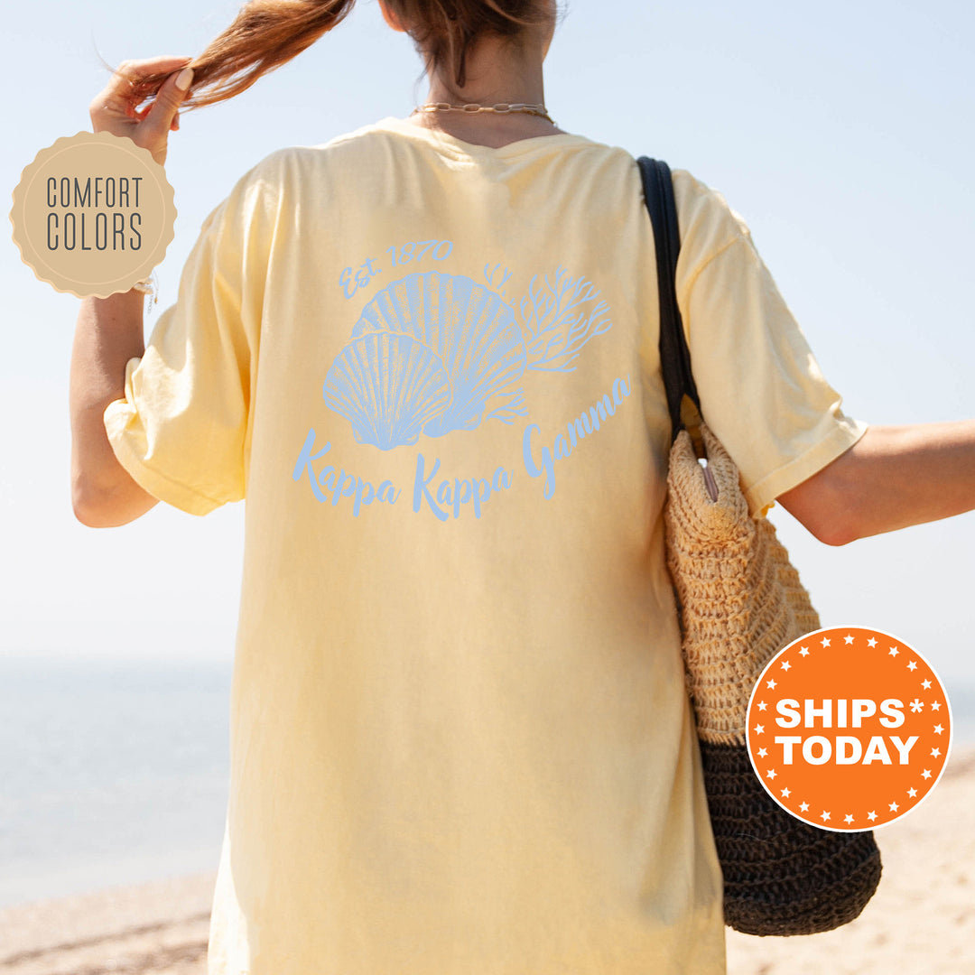 A woman wearing a yellow t-shirt with a blue design and text, standing on a beach with her hair in a ponytail.