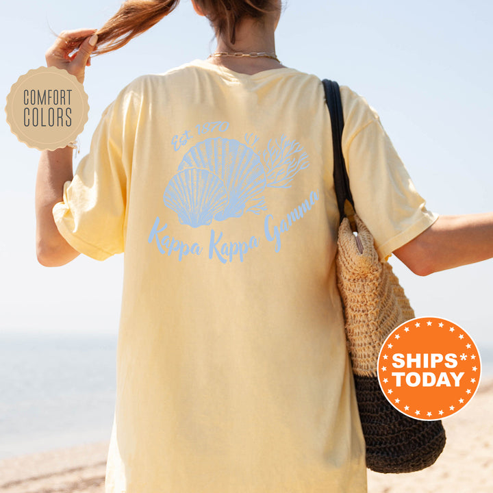 A woman wearing a yellow t-shirt with a blue design and text, standing on a beach with her hair in a ponytail.