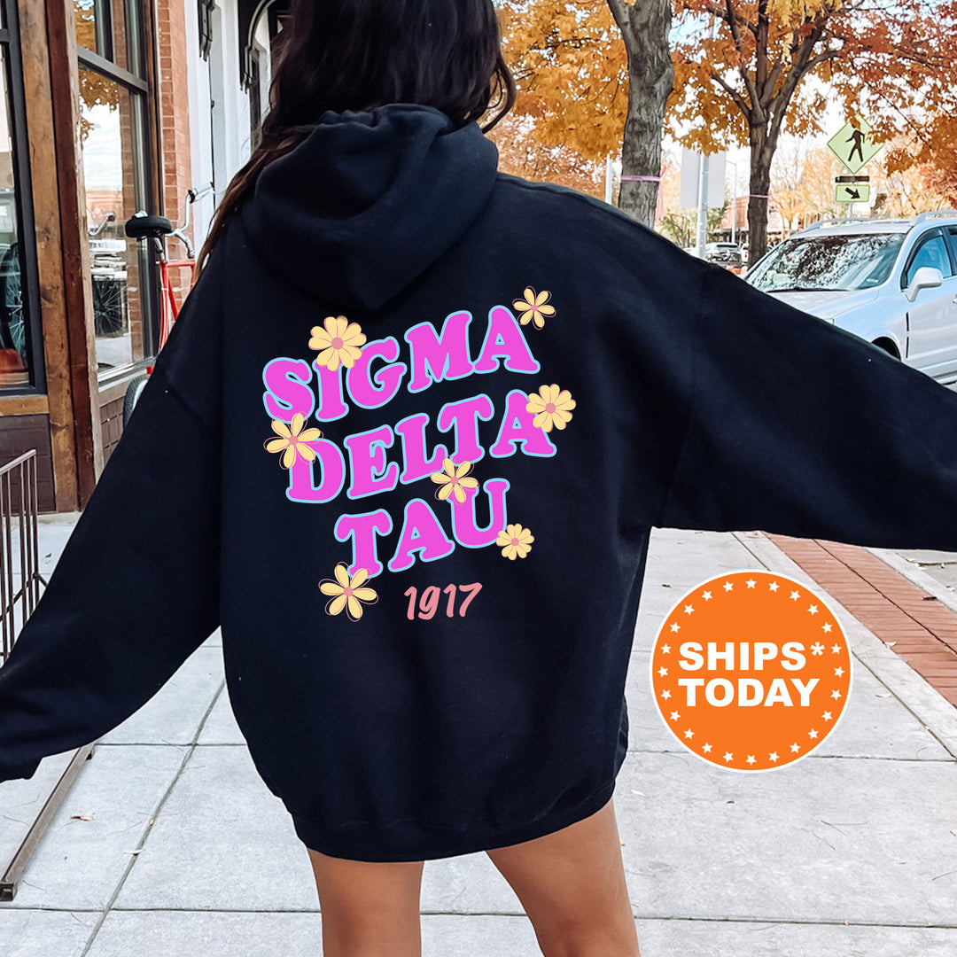 A person wearing a black hoodie with the text "SIGMA DELTA TAU" printed on it, standing on a sidewalk with a car and a tree in the background.