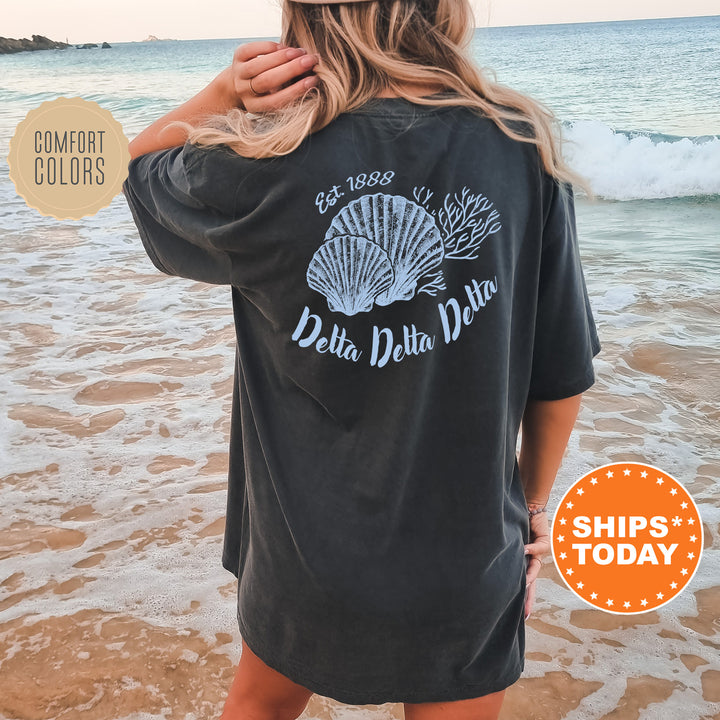 A woman wearing a black t-shirt with a graphic of seashells and the text "Delta Delta Delta" standing on a beach.
