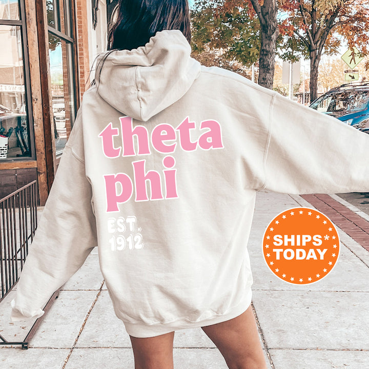 A woman wearing a white hoodie with the text "theta phi" printed on it, standing on a sidewalk.