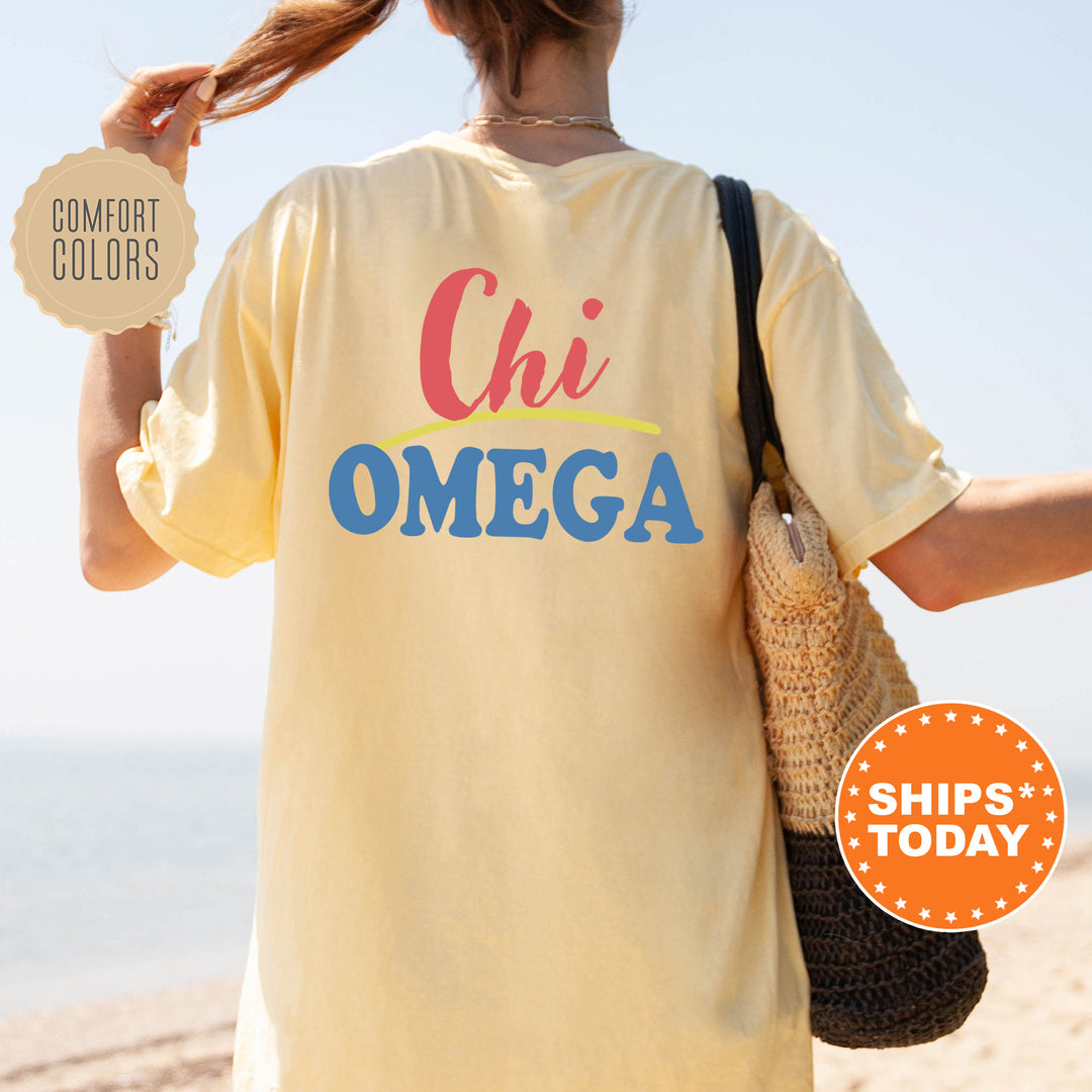 A person wearing a yellow t-shirt with the text "Chi Omega" printed on it, standing on a beach with their hair in a ponytail.