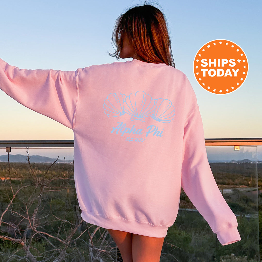 A woman wearing a pink sweatshirt with a graphic design of shells on it, standing in front of a scenic landscape with mountains and a clear sky.