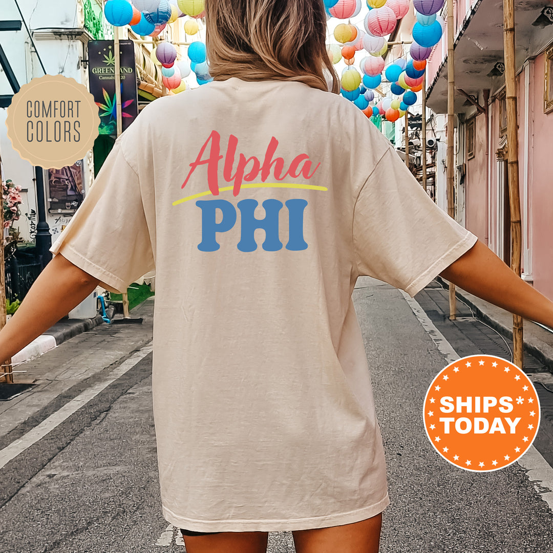 A person wearing a white t-shirt with the text "Alpha Phi" printed on it, standing on a street with colorful lanterns hanging overhead.